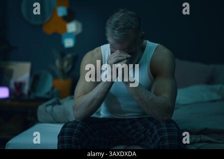 Middle-aged man sitting on a couch in a dimly lit room with his head in his hands, deep in thought. Various household items and furniture in surroundi Stock Photo