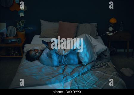 Person lying on bed while looking at smartphone in cozy nighttime room featuring bedside table with lamp, books, and plants around, setting a peaceful Stock Photo
