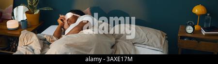 African American man lying in bed, sleeping peacefully in cozy bedroom. Soft lighting enhancing calm and relaxed atmosphere with various items on beds Stock Photo