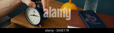 Close-up of hand adjusting alarm clock on bedside table while another hand uses smartphone sleep tracker app beside glass of water and lamp Stock Photo