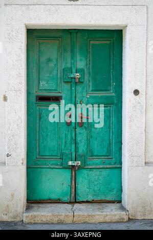A weathered, teal-colored double door is framed by a textured white wall. The door features a mail slot, ornate handles, and is secured with a padlock Stock Photo