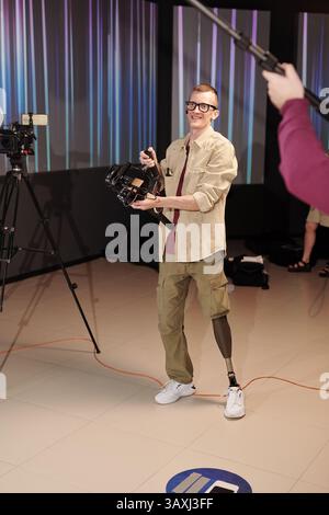Young cheerful cameraman with leg prosthesis working in studio Stock Photo