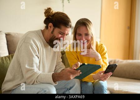 Couple laughing and using tablet and smartphone on sofa at home Stock Photo