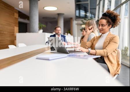 Businesswoman wearing eyeglasses while analyzing data over laptop in office with coworkers in the background Stock Photo