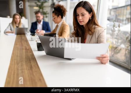 Focused accountant holding report and working over laptop while teammates collaborating in the background Stock Photo