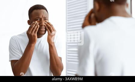 Concerned African Man Touching Bags Under Eyes Standing In Bathroom Stock Photo