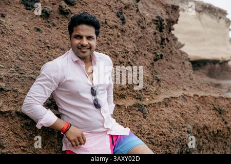 A confident smiling Indian man posing outdoor leaning on a rock at the beach. Stock Photo