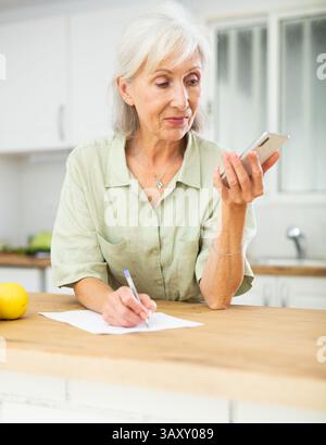 Elderly woman using phone and writing notes Stock Photo