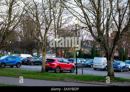 A parking lot in Saffron Walden, Essex, filled with various cars and surrounded by trees. The scene is illuminated by street lights under a cloudy sky Stock Photo
