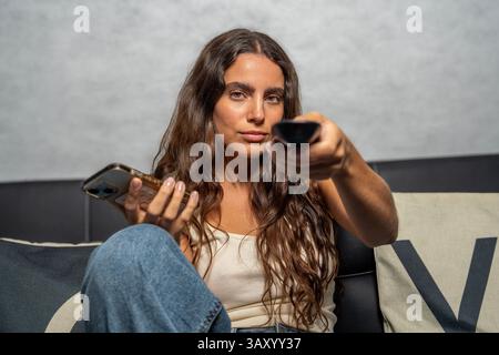 Young woman sitting on sofa using smartphone and holding remote control, watching television Stock Photo