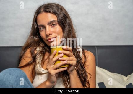 Serene young woman enjoying a quiet moment on the sofa, cradling a warm mug in her hands Stock Photo