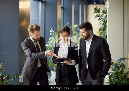 Business professionals standing in office hallway, discussing project and analyzing data on tablet. Stock Photo