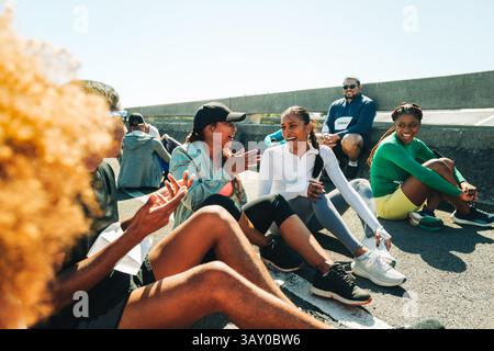 A group of friends sit and relax on the ground after finishing a marathon. They are engaging in lively conversation and laughter, with warm sunlight a Stock Photo