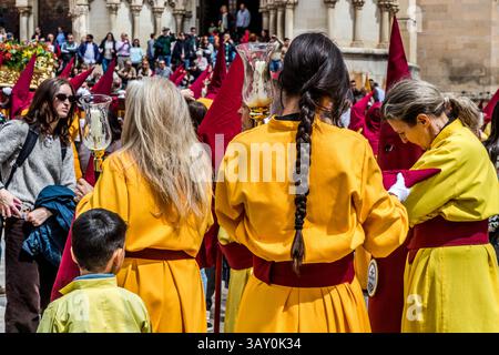 During a break in the Good Friday procession Camino del Calvario on the Plaza Mayor of Cuenca, brotherhood members otherwise hidden beneath their pointed hoods (capirotes) can occasionally be recognised as individuals, Castilla-La Mancha, Spain. Two women and a girl in yellow robes with hoods pushed back carry lantern staffs, while hooded penitents remain in the background. During Semana Santa in Cuenca, 33 Hermandades take part in eleven processions through the city during Holy Week. Stock Photo