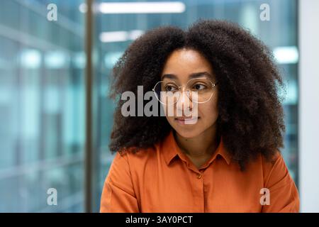 A thoughtful young woman with curly hair and glasses looks off to the side, in a modern office setting. Her orange blouse complements her warm complexion. Stock Photo
