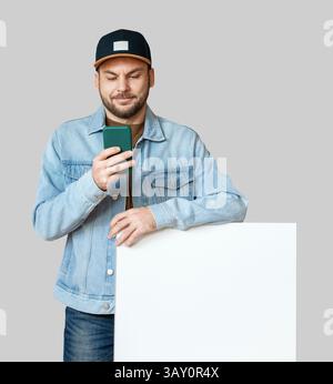 Isolated young adult man in casual outfit and snapback using smartphone while leaning on white blank poster. Stock Photo
