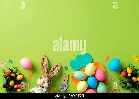 Happy Easter concept. Flat lay bunny, colorful Easter eggs in a carrot-shaped plate, and Easter decorations on green background. Stock Photo