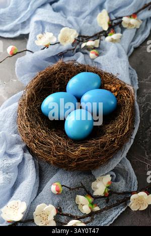 Bright blue Easter eggs nestled in a bird's nest, surrounded by white blossoms and a blue cloth on a textured stone background. Stock Photo