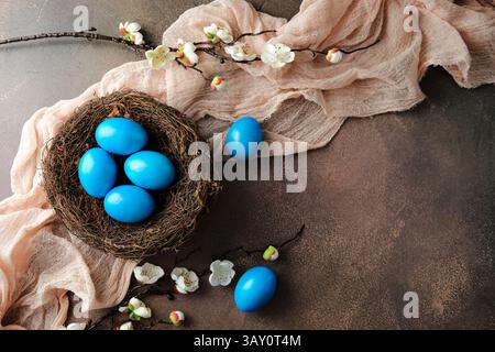 Bird's nest with blue Easter eggs, accented by white cherry blossom branches and soft beige fabric on brown textured background. Happy Easter greeting Stock Photo