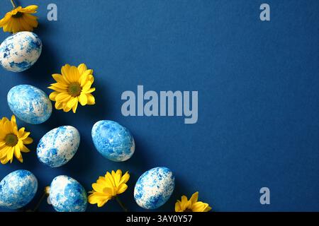 Happy Easter greeting card with painted eggs and yellow flowers on dark blue background. Top view, flat lay, copy space. Stock Photo