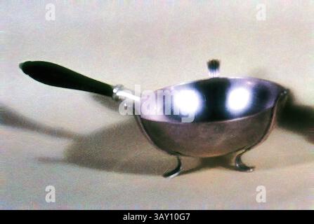 A shiny, elegant saucepan with a curved body and a sleek black handle, resting on three delicate feet. The light reflects off its surface, highlighting its polished finish., Adrian Bancker, American, 1703-1772, Pipe Warmer, ca. 1735, silver and wood, Overall: 2 7/8 × 10 1/2 × 5 1/8 inches (7.3 × 26.7 × 13 cm), Classic art with a modern twist reimagined by Artotop Stock Photo