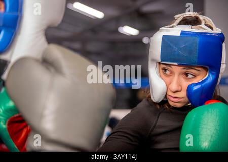Female boxer training, wearing protective headgear and gloves, practicing punches in a boxing ring, focused on her opponent Stock Photo