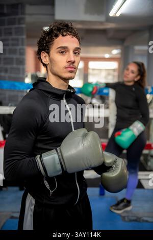 Young hispanic male boxer wearing boxing gloves posing in a gym ring with female boxer in background Stock Photo