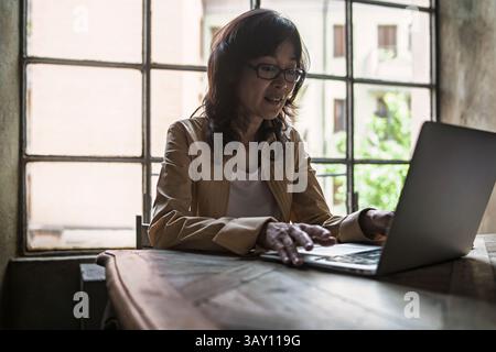 Mature asian woman using laptop by large window in elegant workspace with soft daylight and green plant background Stock Photo