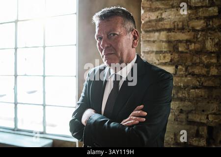 Confident senior businessman in elegant suit standing near bright window in stylish office, looking assertive and authoritative Stock Photo