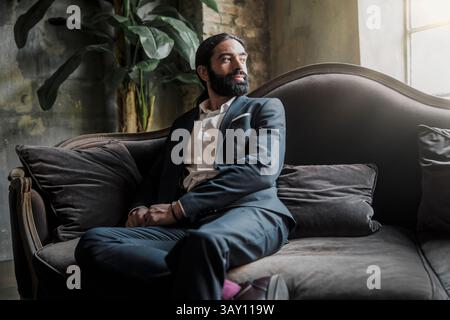 Stylish hispanic man in suit sitting on classic velvet sofa in dramatic natural light, looking away thoughtfully Stock Photo