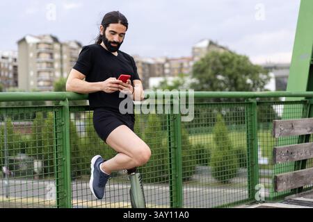 Active man with prosthetic leg leaning on urban bridge rail while texting on smartphone, modern inclusive lifestyle and mobility Stock Photo
