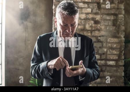 Confident senior businessman checking his smartphone while standing near window in elegant office space with rustic wall and sunlight Stock Photo