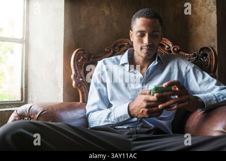 Relaxed young man using smartphone while sitting on vintage leather armchair by window. Casual indoor setting with natural light. Stock Photo