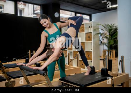 Pilates instructor helping a student performing an advanced stretching exercise on a reformer machine in a gym Stock Photo