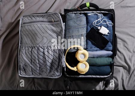 carry-on suitcase with blue clothes and large headphones, charger and cell phone on the bed Stock Photo