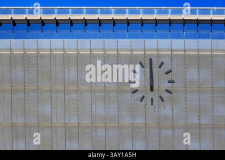 Minimalist wall clock with black dial and hands on the facade of a modern building with blue glass and fine metal mesh, captured in daylight Stock Photo