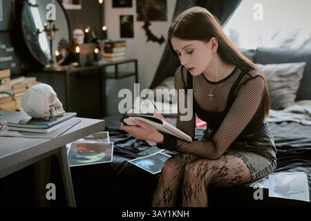 Brunette goth teen in black clothes with dark make up and cherry lips drawing in notepad while sitting on bed in her room Stock Photo