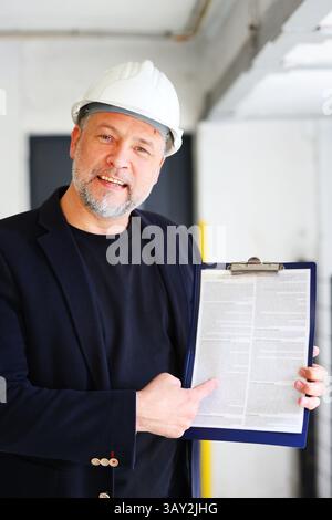 Smiling construction manager in hard hat pointing at contract on clipboard, presenting documentation indoors. Copy space, moc up Stock Photo