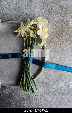 From above, white daffodils with a vibrant blue ribbon on a textured grey surface. Stock Photo