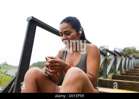 A young ecuadorian woman sits on outdoor steps, enjoying music or a podcast through wireless earbuds while using her smartphone. She appears relaxed, Stock Photo