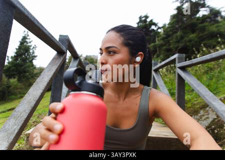 An Ecuadorian woman sits outdoors holding a red water bottle, appearing focused and thoughtful. Surrounded by nature in Loja, Ecuador, she wears wirel Stock Photo