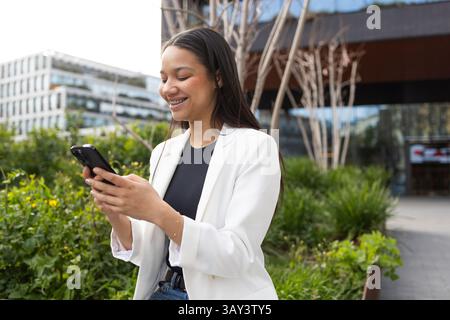 Smiling Hispanic woman in white blazer using smartphone outside a modern office building. Represents business lifestyle, connectivity, and technology. Stock Photo