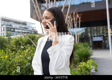 A confident hispanic businesswoman is standing outdoors, talking on her smartphone. She wears a white blazer, exuding a professional and dynamic busin Stock Photo