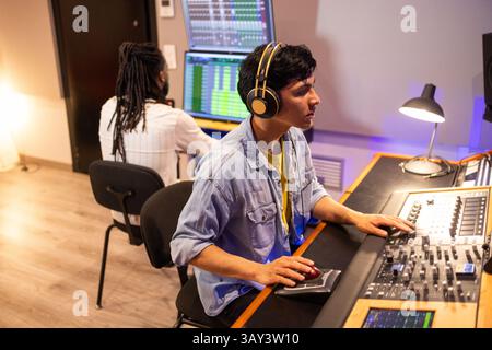 Two audio engineers work in a recording studio, wearing headphones and using mixing consoles and monitors to edit and produce music tracks. Focused, c Stock Photo