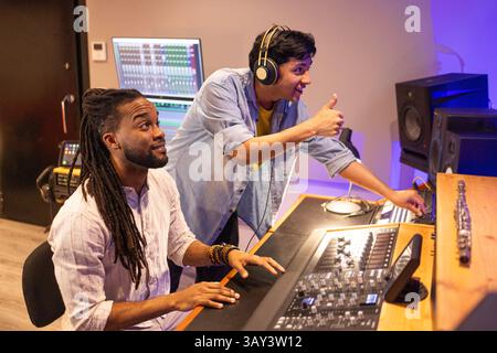 A vibrant recording studio scene showing two focused producers working on audio mixing. One uses a mixing console while the other gestures positively Stock Photo