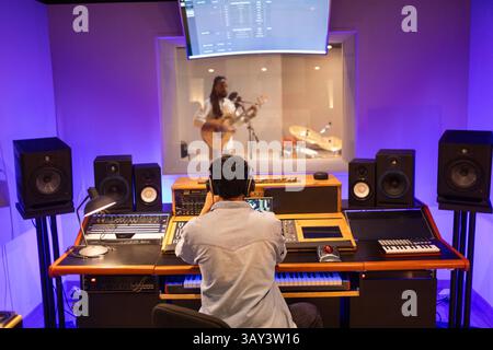 Musician plays guitar behind soundproof glass while engineer operates mixing console. Studio filled with advanced equipment, creating a dynamic record Stock Photo