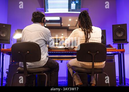 Two audio engineers in a modern recording studio focusing on music production. They sit at a mixing console with monitors, showcasing a professional s Stock Photo