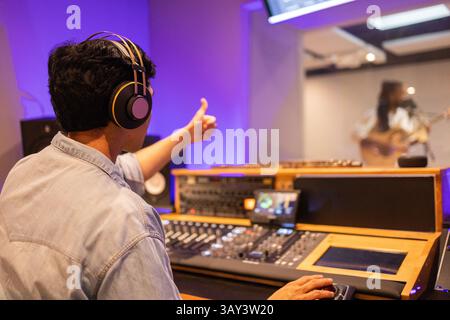 A skilled sound engineer, wearing headphones, gives a thumbs-up in a modern recording studio. He oversees a session from a high-tech soundboard as a m Stock Photo