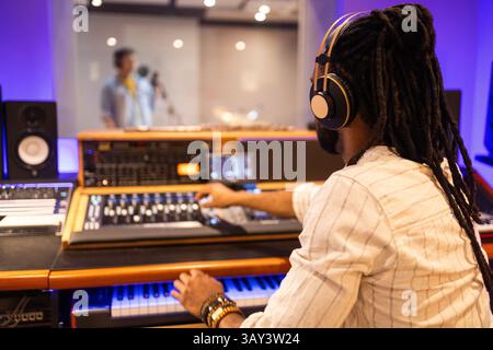 A music producer with rasta dreadlocks works at a professional mixing console while wearing headphones. Through the glass, a vocalist performs in the Stock Photo