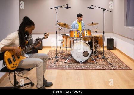 Two musicians collaborating in a modern recording studio. One plays drums, and the other plays bass guitar. The room has soundproofing and microphones Stock Photo